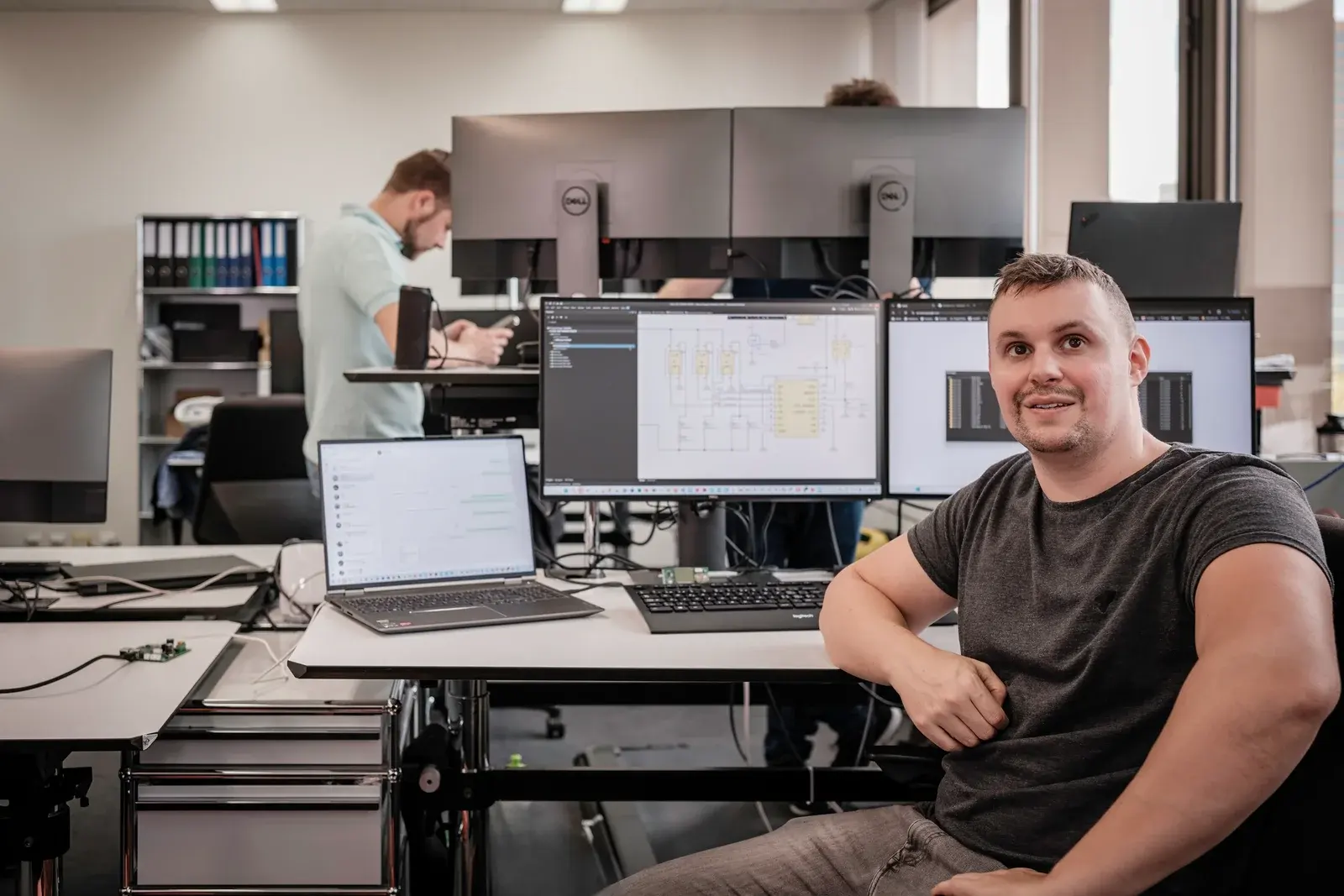 Akrodyn-Entwickler an seinem Arbeitsplatz mit Schaltplan und Terminal auf den Bildschirmen